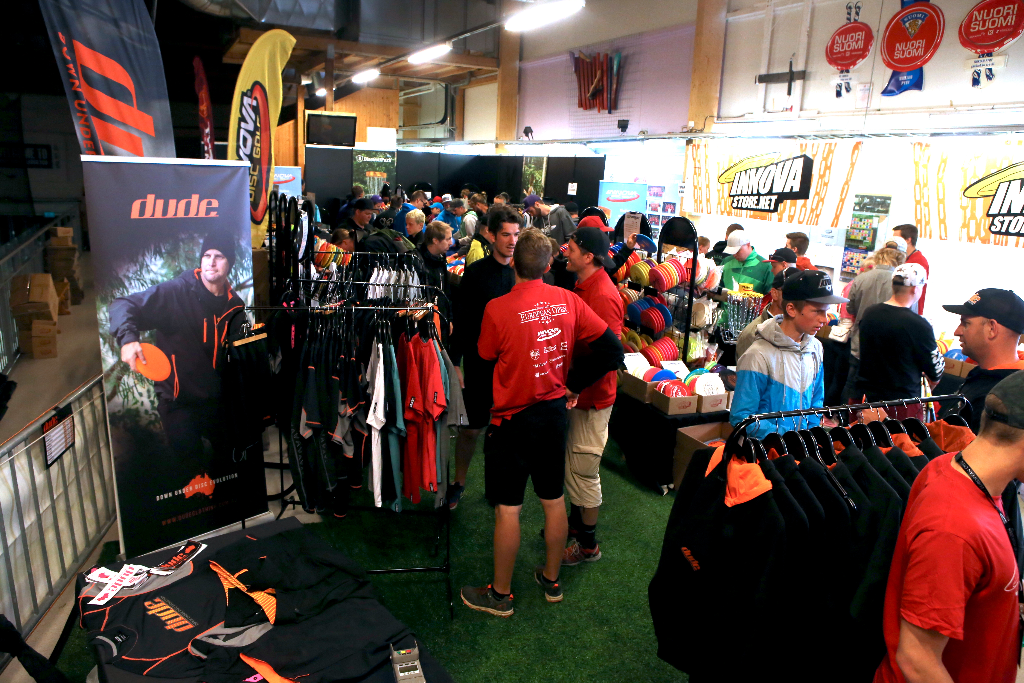 A busy disc golf trade stand at an expo, with racks of clothing, discs and a crowd of visitors.
