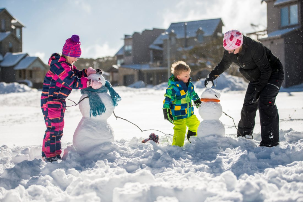 Children and an adult building a snowman outside alpine village lodges.