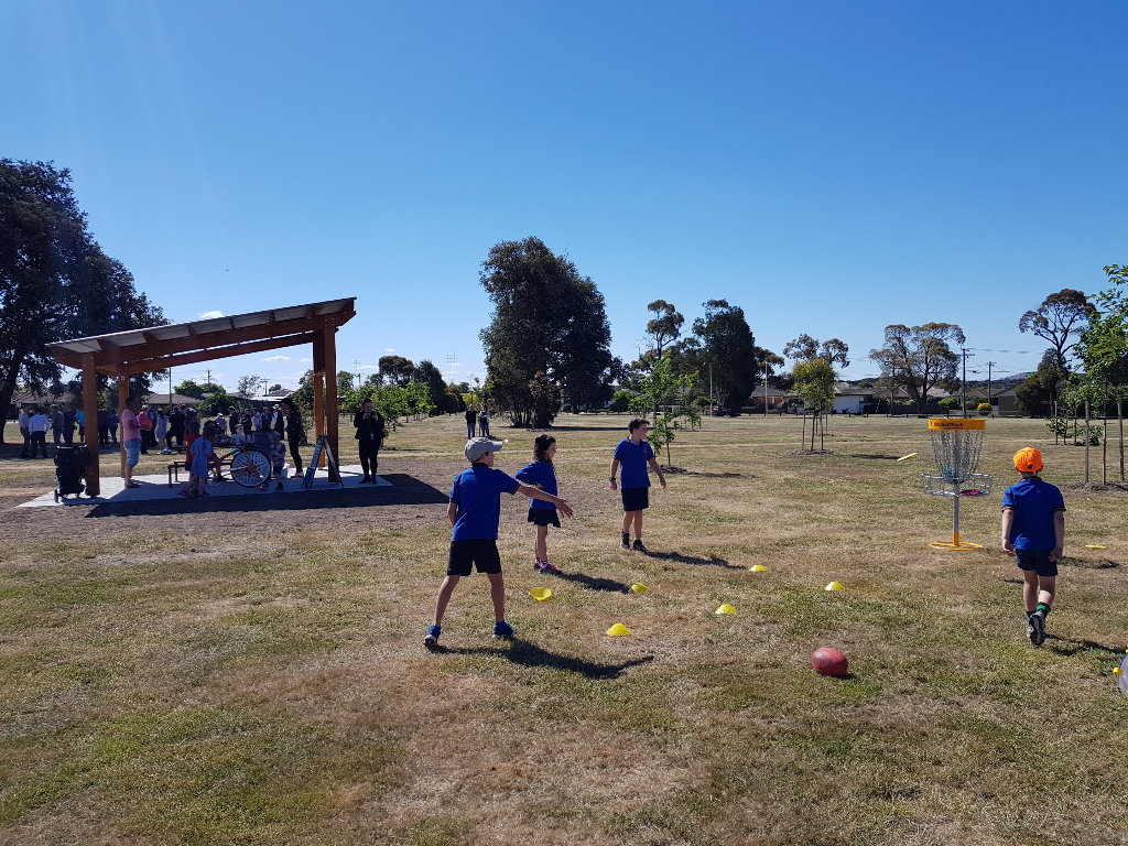 School children playing around a disc golf basket on a school oval beside a shade shelter.