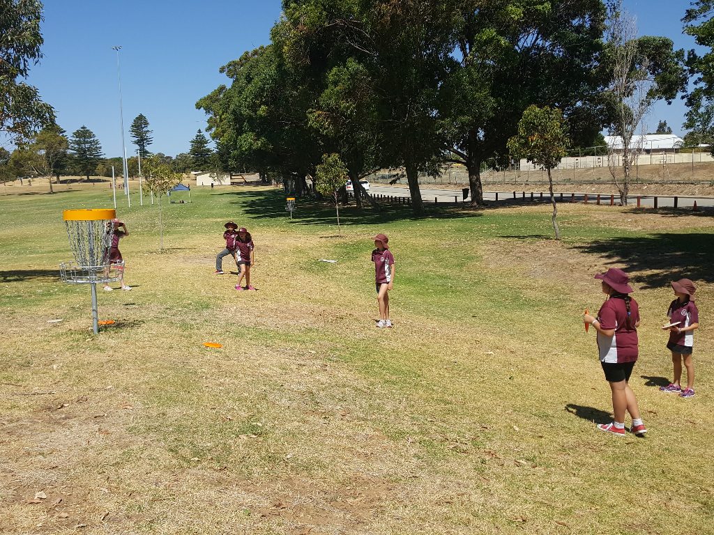 Children playing around a disc golf basket on dry grass shaded by gum trees.
