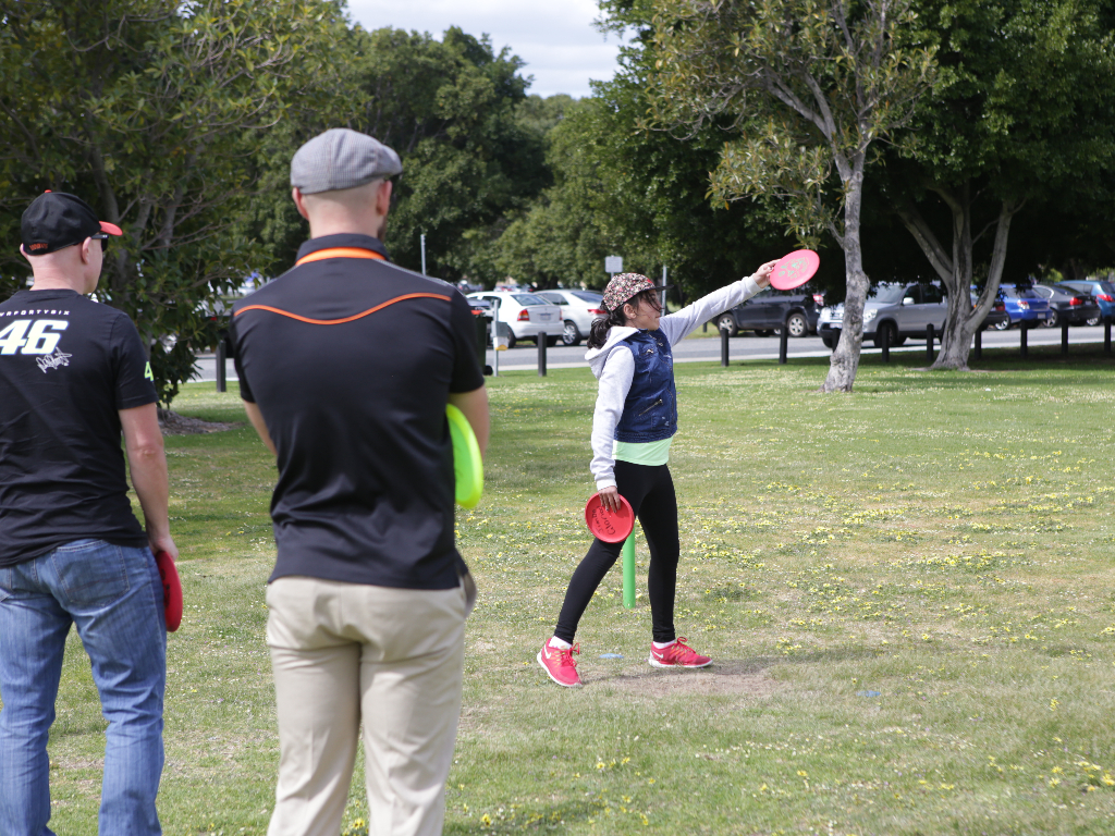 A girl throwing a disc on grass while two men watch, cars parked behind.