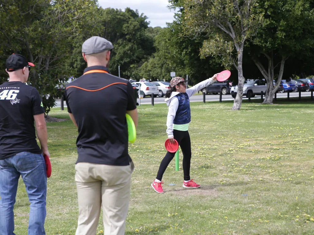 A girl throwing a disc on grass while two men watch, cars parked behind.