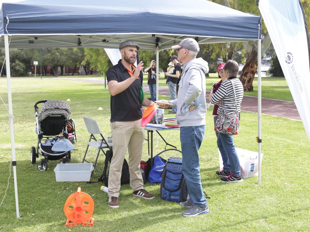 People talking under a marquee at a come-and-try day, equipment and a pram beside them on the grass.