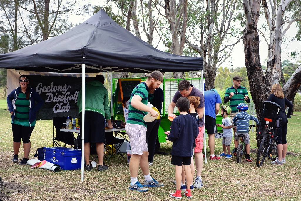 Families and children gathered at a Geelong Disc Golf Club marquee, bikes parked alongside.