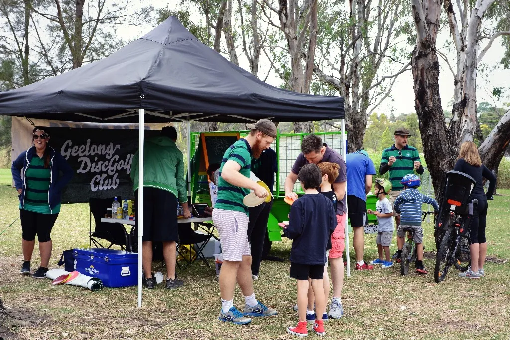 Families and children gathered at a Geelong Disc Golf Club marquee, bikes parked alongside.