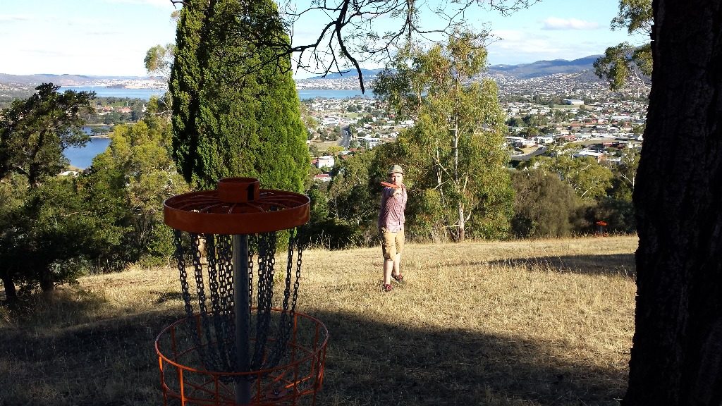 A disc golf basket on a dry hillside overlooking a town and river valley.