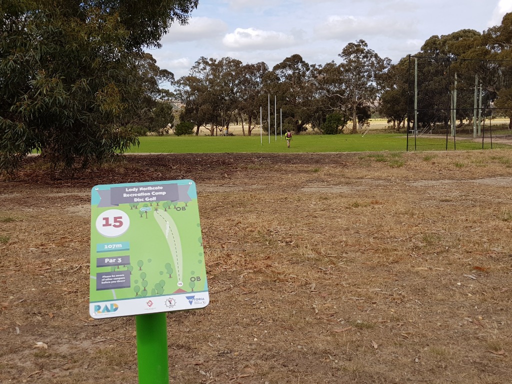 A tee sign for hole 15, par 3, on a green post beside a bare dirt tee area.
