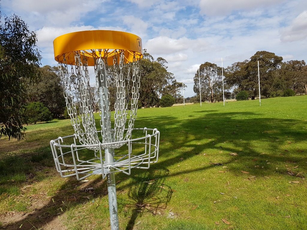 A yellow-topped disc golf basket on green grass beside a sports field.