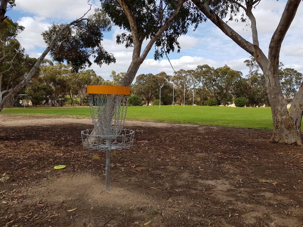 An orange-topped disc golf basket on bare ground beneath gum trees.