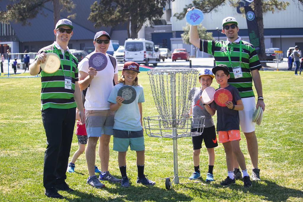 Adults and children posing with discs around a basket at a festival of sport, city buildings behind.