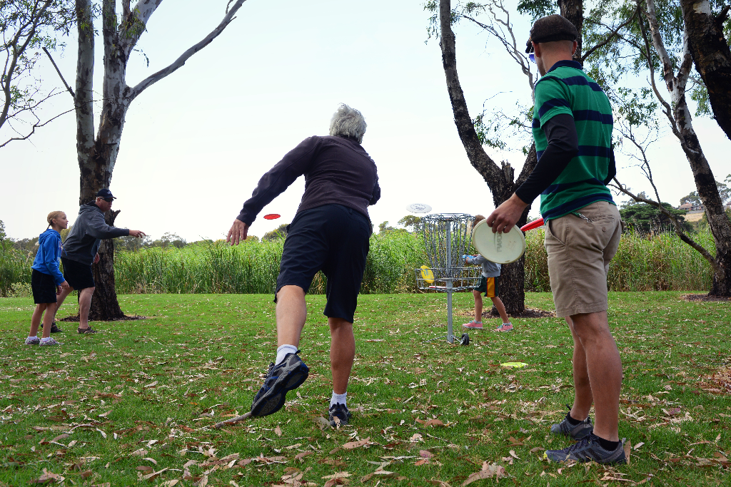 An older woman throwing a disc on grass while a man watches from beside the tee.