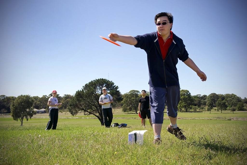 A player throwing an orange disc across open parkland, others waiting behind.