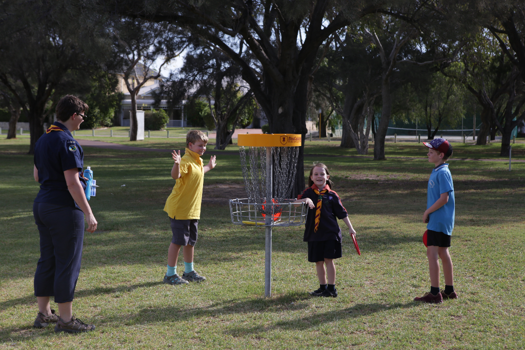 A leader and children in scout uniforms gathered around a disc golf basket on grass.