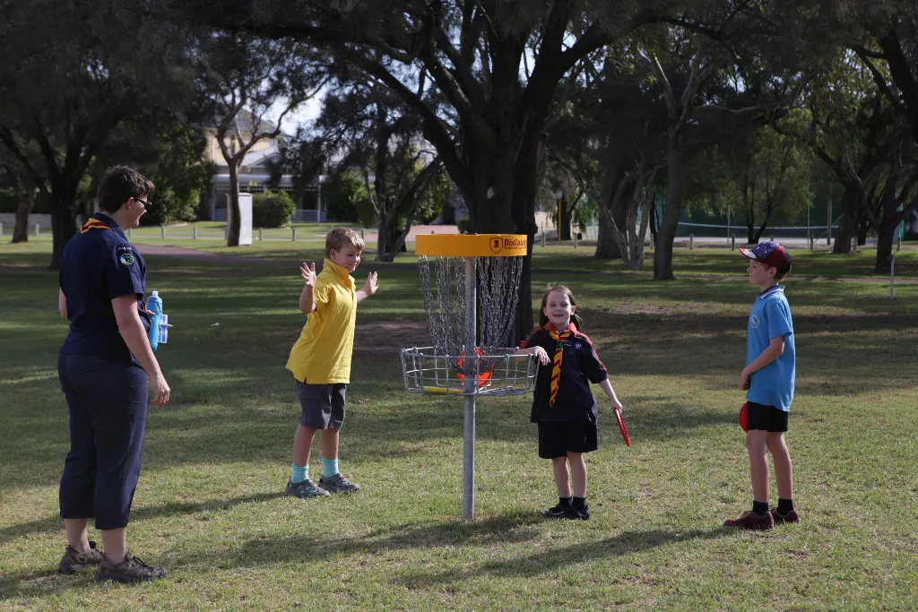 A leader and children in scout uniforms gathered around a disc golf basket on grass.