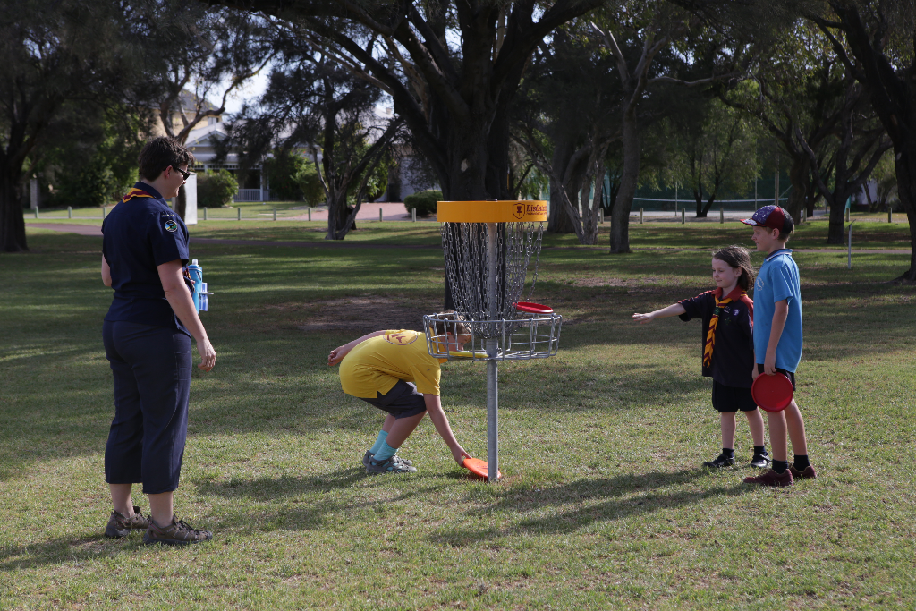 Children around a disc golf basket, one bending to pick up a disc.