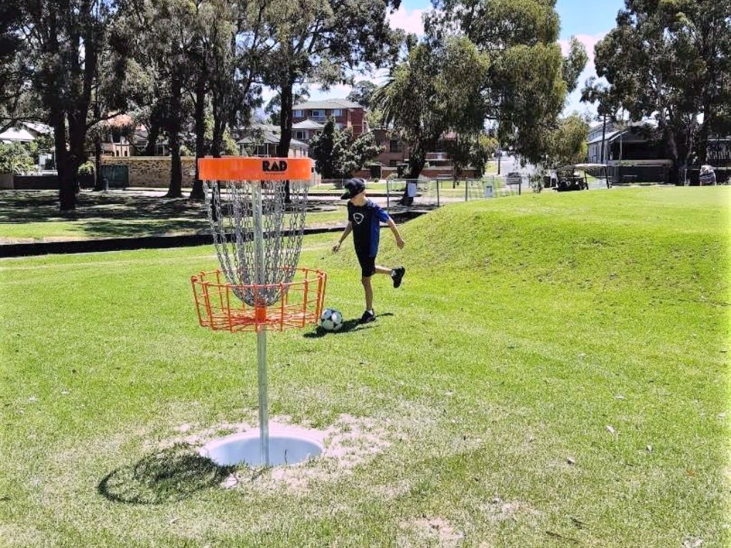 A boy kicking a football towards an orange-topped target basket on a grassy slope.