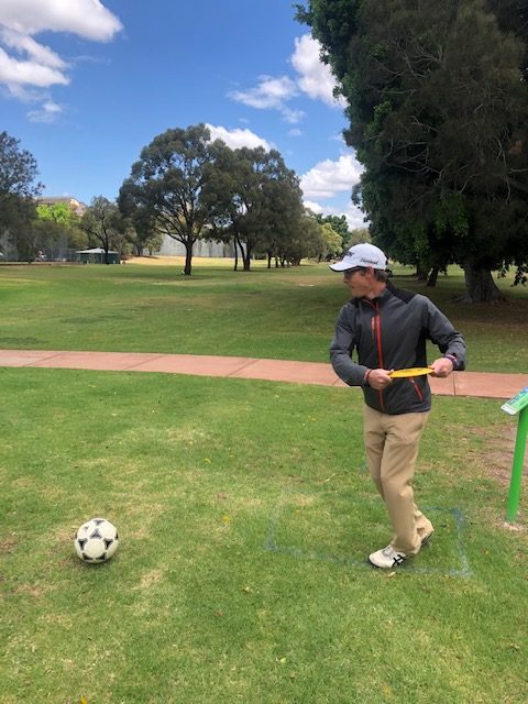 A player walking a fairway carrying a yellow disc, a football resting on the grass nearby.