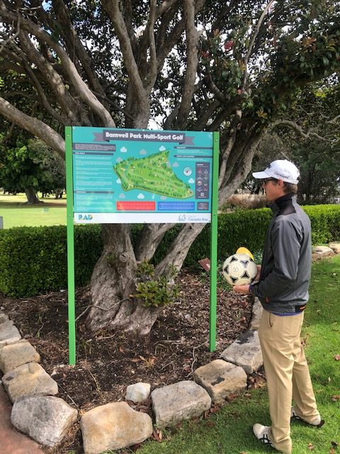 A player holding a football beside the Barnwell Park Multi-Sport Golf information board.
