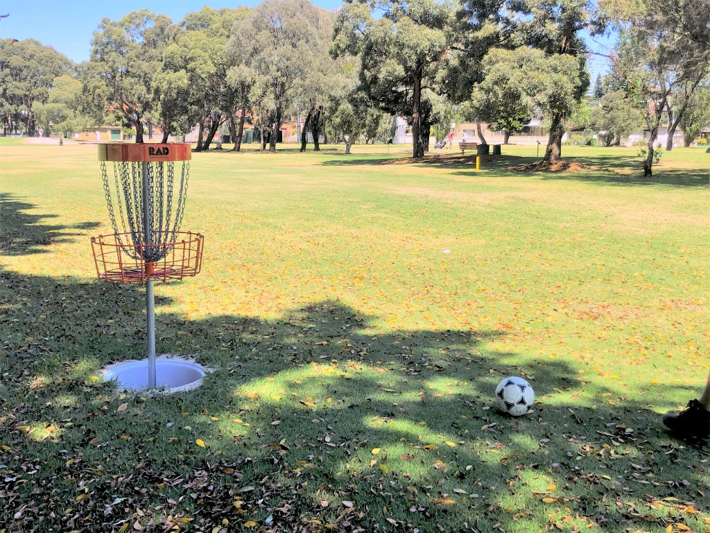 A disc golf basket on a tree-lined fairway with a football on the grass in front of it.