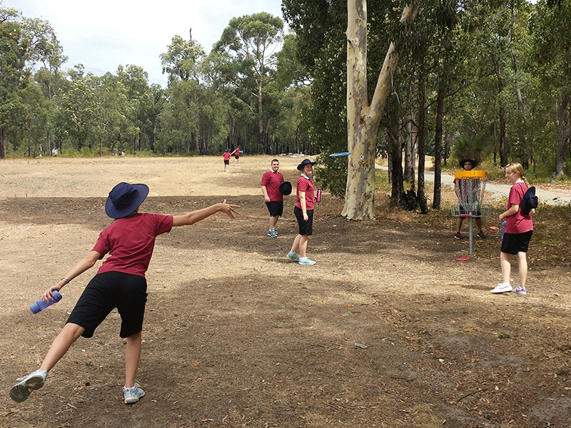 A player in a wide-brimmed hat throwing a disc while children wait their turn on a dry course.