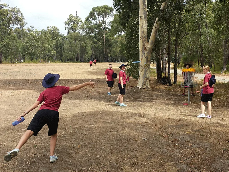A player in a wide-brimmed hat throwing a disc while children wait their turn on a dry course.