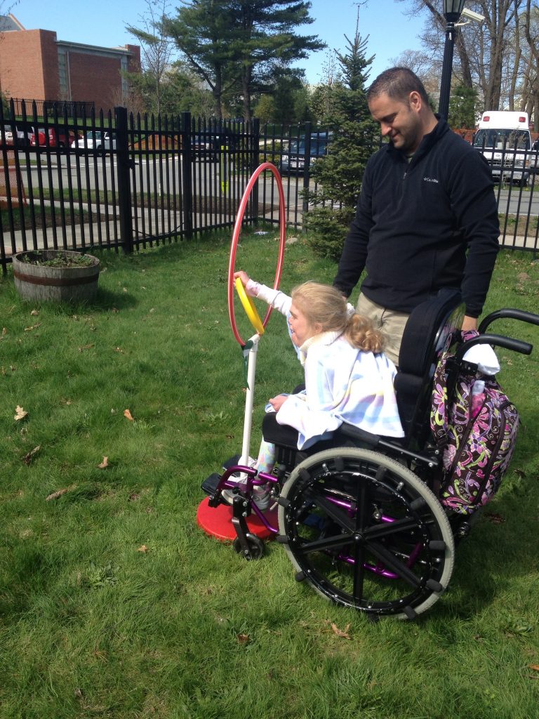 A child in a wheelchair using an adapted launcher to throw, guided by an instructor on a grassy field.
