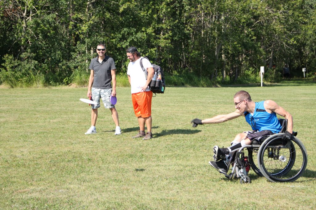 A player in a wheelchair throwing a disc across a grassy course while others watch.
