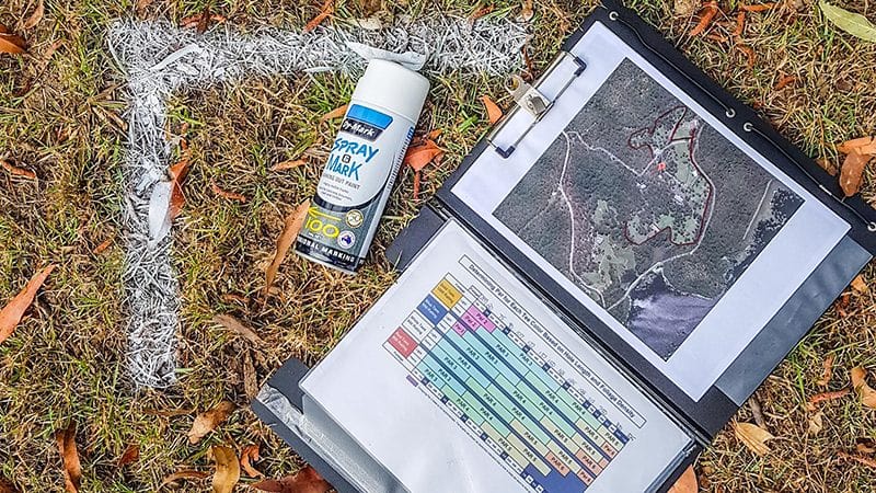 A course design kit on the ground: marking spray and a clipboard holding an aerial site map and hole schedule.