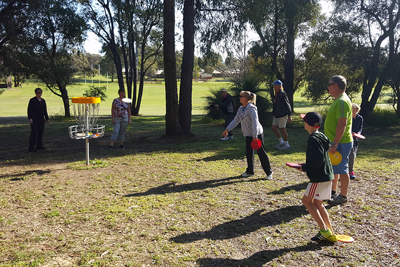 People playing disc golf around a basket in a park, children holding coloured discs.