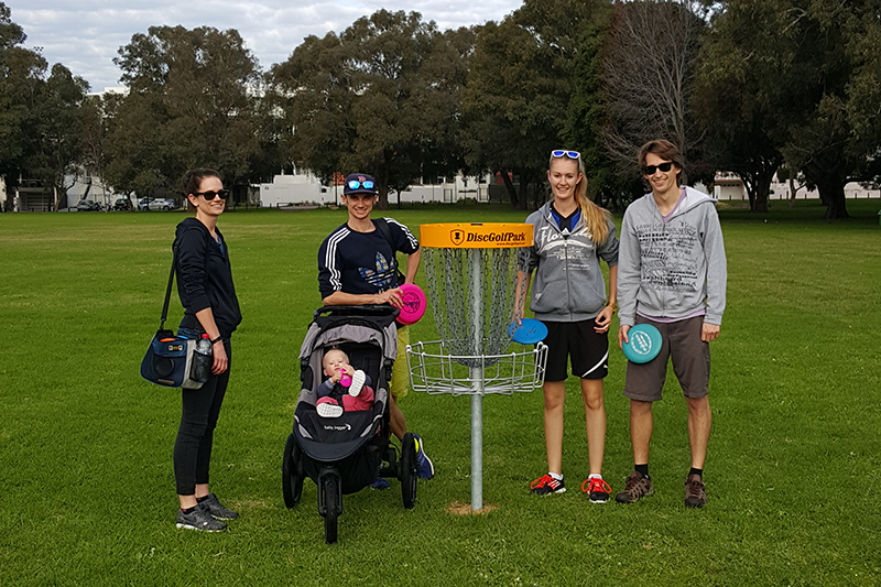 A group posing beside a disc golf basket with discs and a pram.