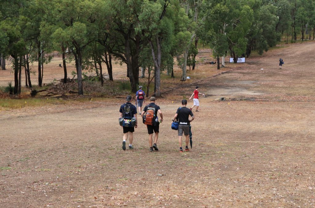 Players walking a dirt track through bushland carrying disc golf bags.