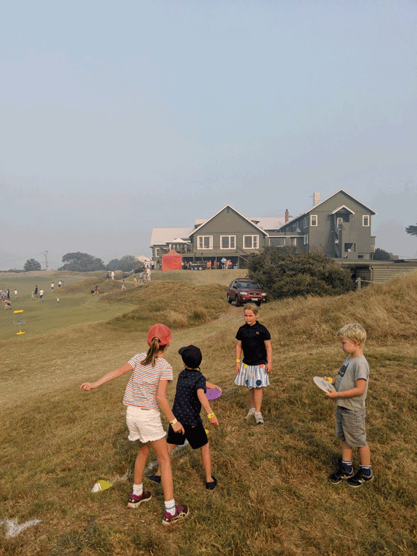 Children carrying discs across a grassy dune, the clubhouse on the ridge behind.
