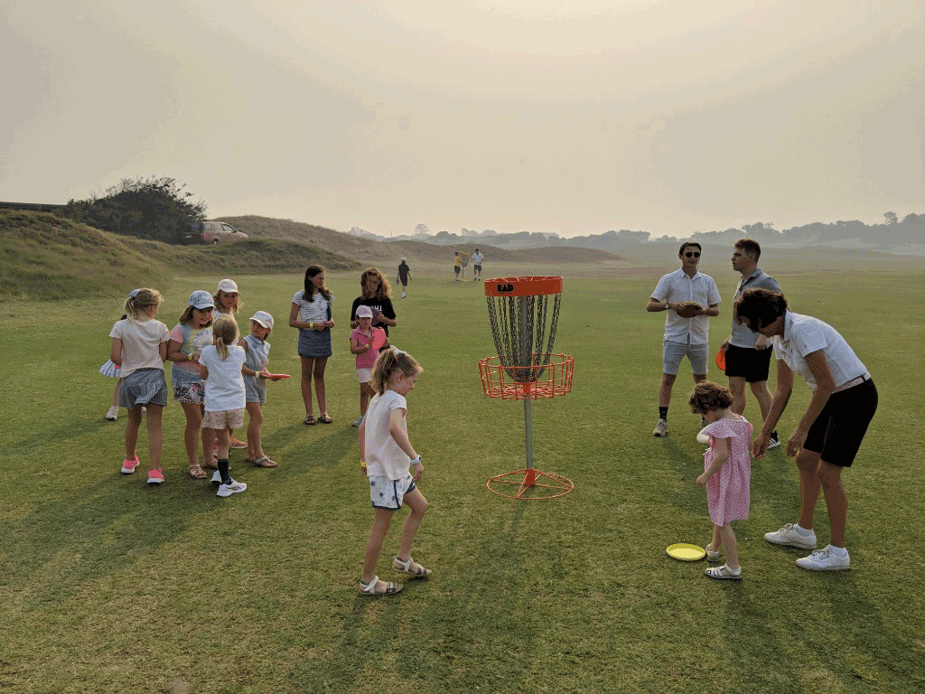 Children and adults gathered around an orange basket on links-style dunes in hazy light.