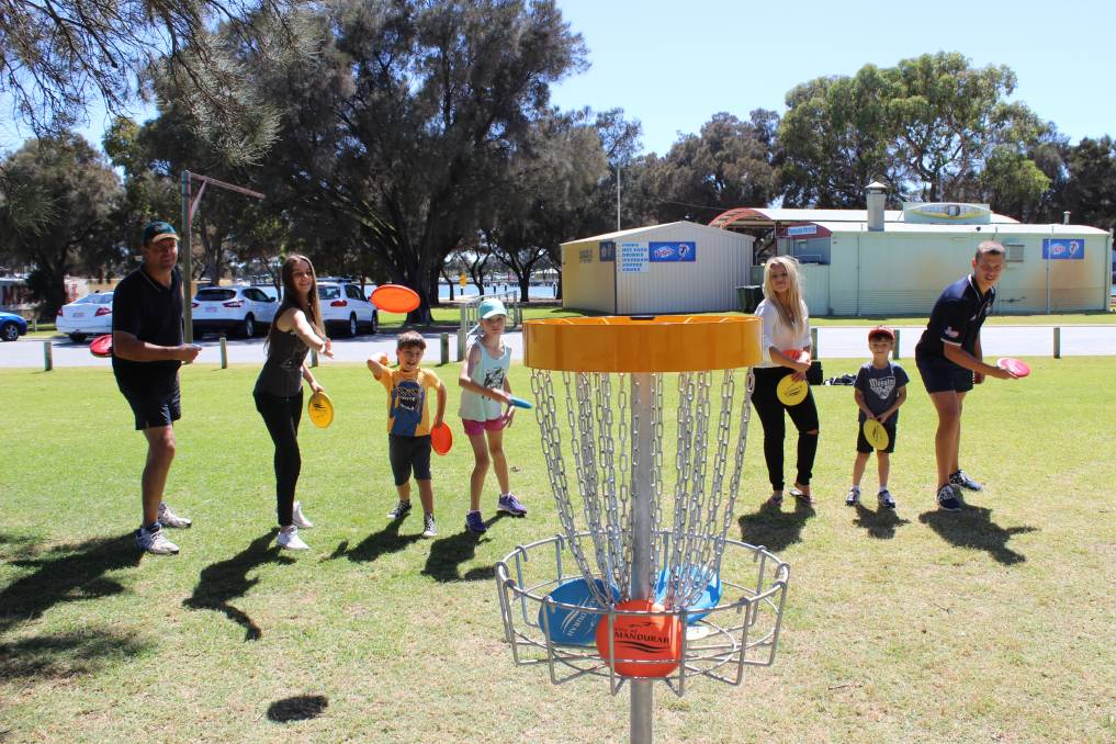 Adults and children gathered around an orange-topped basket on grass, caravans parked behind.
