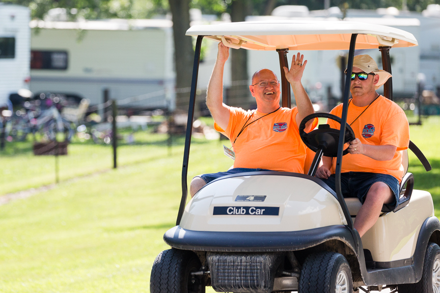 Two event volunteers in orange shirts driving a golf buggy across a course.