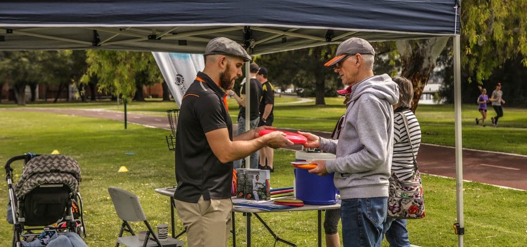 A man handing out discs to visitors at a marquee equipment table.