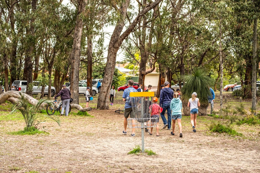 Families playing disc golf around a basket in bushland, bikes and cars parked behind.
