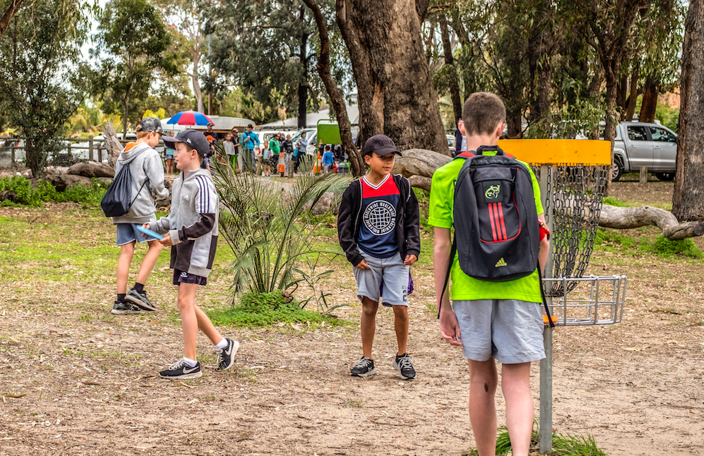 Teenagers with backpacks walking past a disc golf basket on a bushland course.