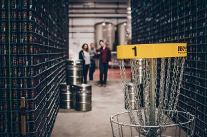 A disc golf basket marked hole 1 set inside a brewery among the fermentation tanks, with drinkers behind.