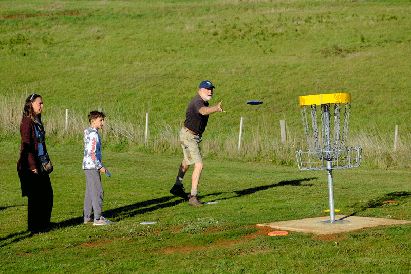 A player throwing towards a yellow-topped basket on a green slope while a woman and boy watch.