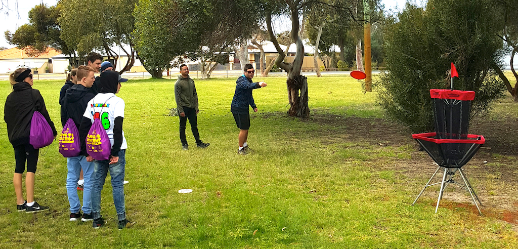 A group putting at a portable disc golf basket on a lawn beside trees.