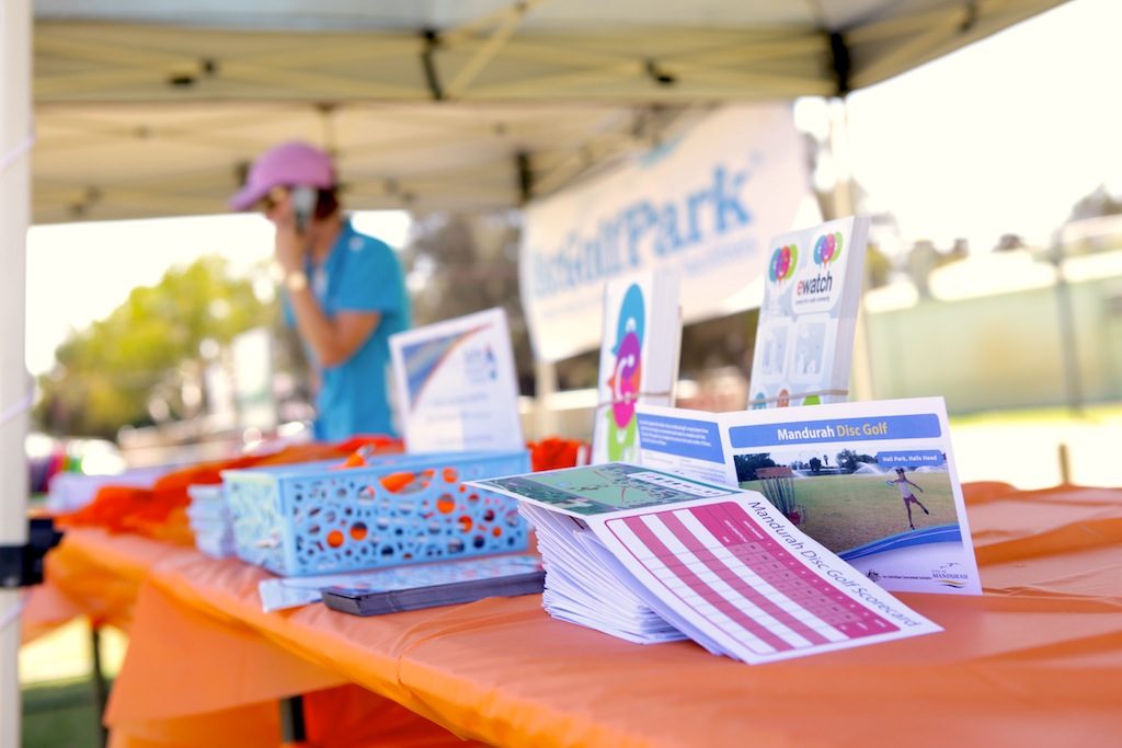 A staff member at an information table under a marquee, course flyers and discs laid out in front.