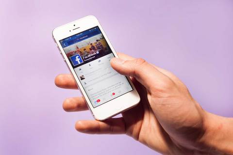 A hand holding a phone showing a Facebook feed.