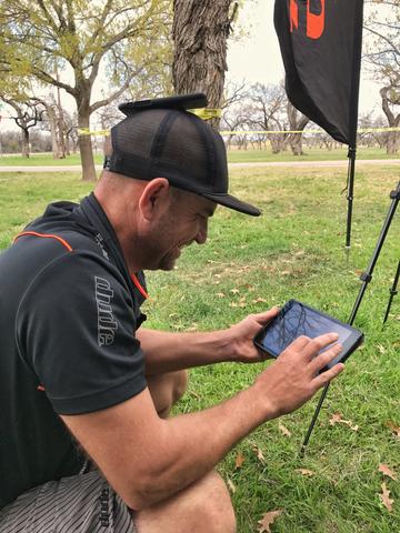 A man kneeling on the grass using a tablet at an event, a feather flag behind him.