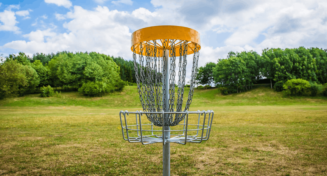 A yellow-topped disc golf basket on green grass with trees behind under a blue sky.