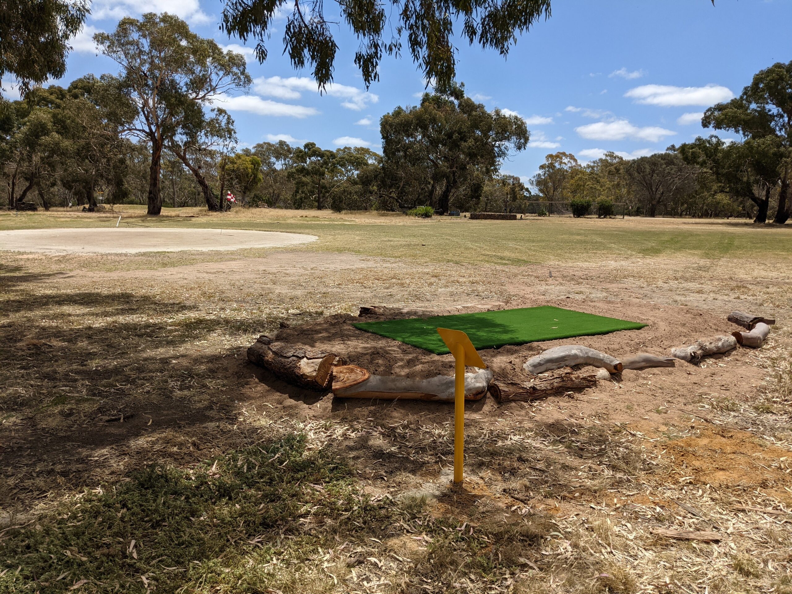 A green artificial tee pad edged with logs, looking out across the fairway to a golf green.