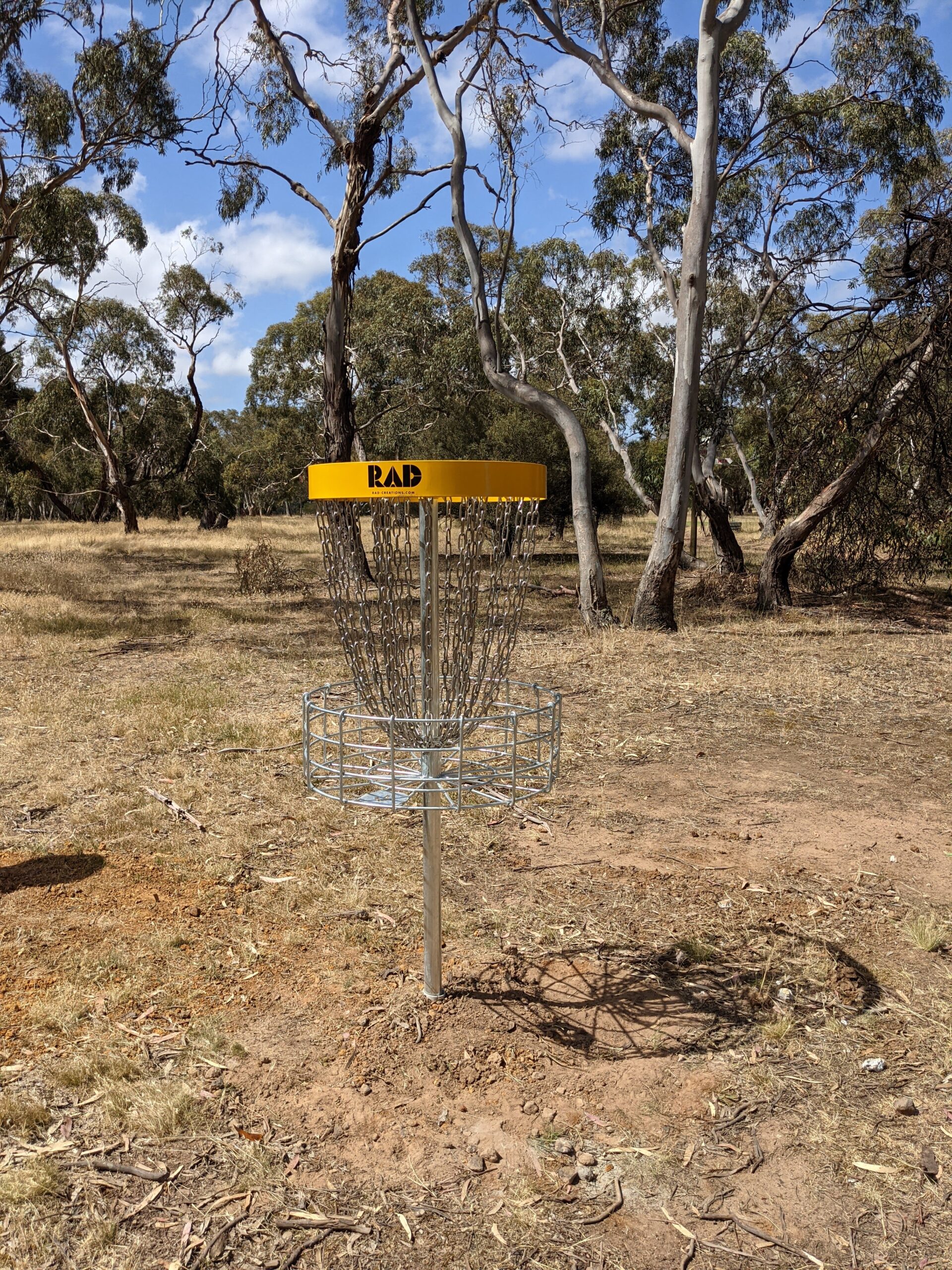 A RAD disc golf basket standing on dry ground among gum trees.