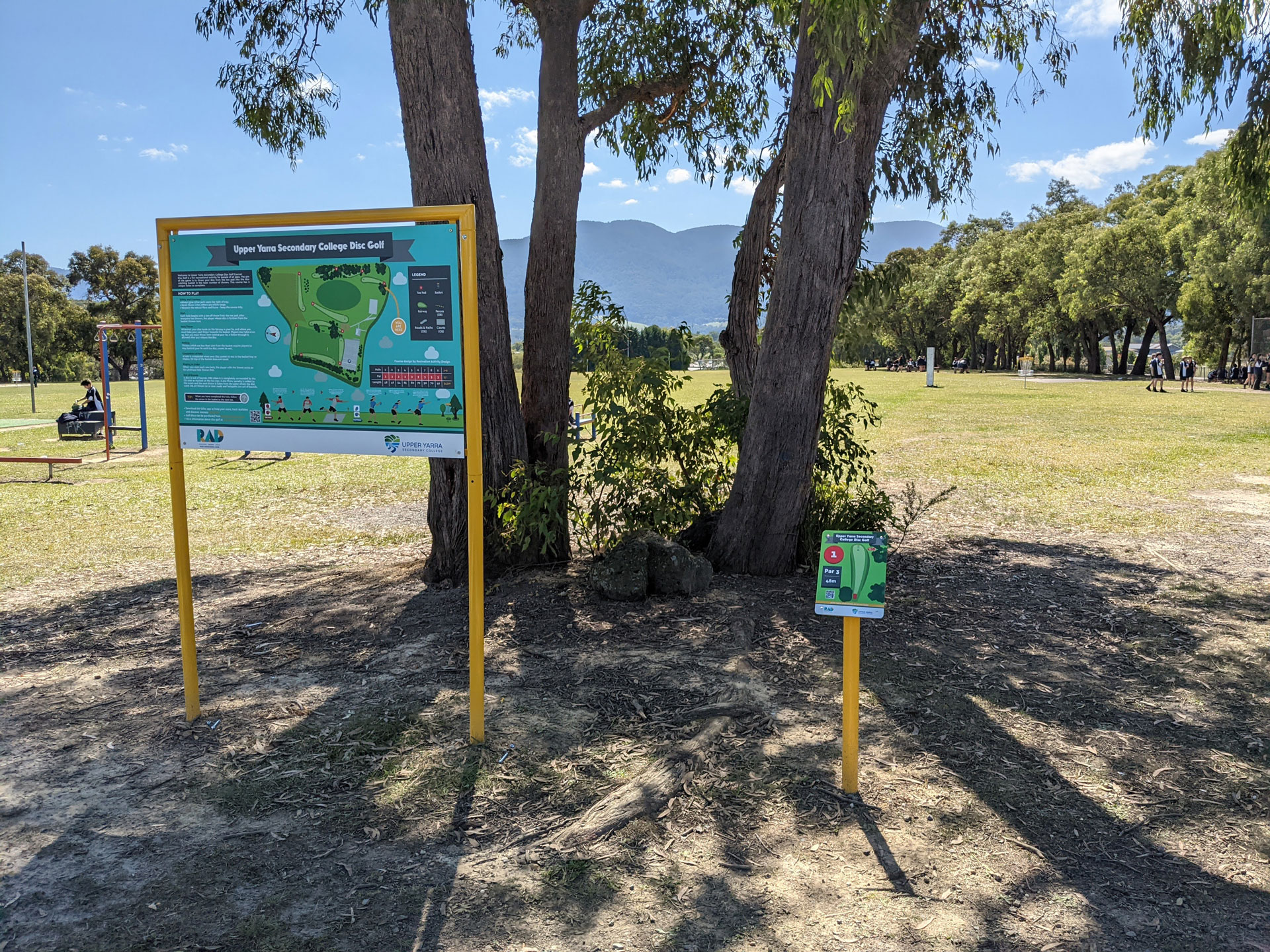 The Upper Yarra Secondary College disc golf information board on yellow posts, with mountains behind.