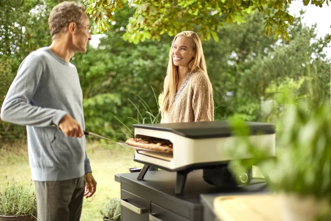 A man and a woman standing next to an outdoor pizza oven.