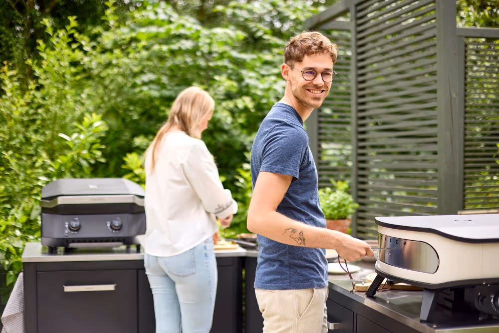 A man standing next to a woman cooking food on top of a grill.